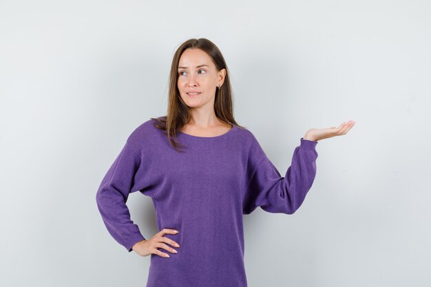 Młoda kobieta w fioletowej koszuli podnosząc dłoń jak coś trzymającego i patrząc zamyślony, widok z przodu.