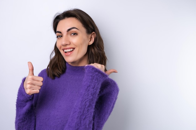Młoda kobieta w fioletowym miękkim, przytulnym swetrze na tle słodkiego uśmiechniętego radośnie w świetnym humorze wskazuje palcem w prawo na pustą przestrzeńx9