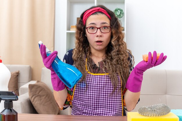 Młoda kobieta w gumowych rękawiczkach trzymająca spray do czyszczenia i gąbkę zaskoczona, siedząc przy stole z środkami czyszczącymi i narzędziami w jasnym salonie