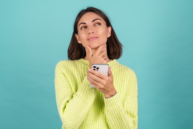 Młoda kobieta w jasnozielonym swetrze z telefonem komórkowym zamyślona i dotykająca jej podbródka