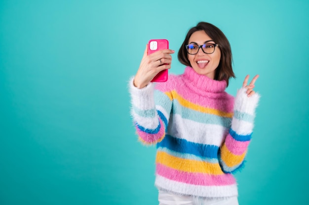 Młoda kobieta w jasnym wielokolorowym swetrze na niebiesko w okularach, trzyma telefon, robi selfie