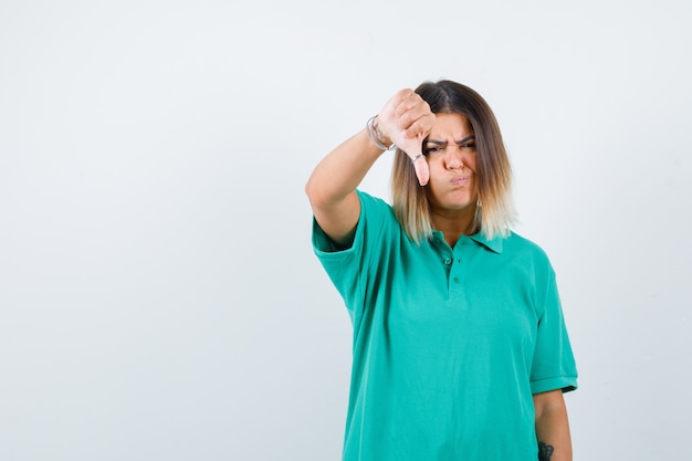 Młoda kobieta w koszulce polo pokazując kciuk w dół z podpuchniętymi policzkami i patrząc niezadowolony, widok z przodu.