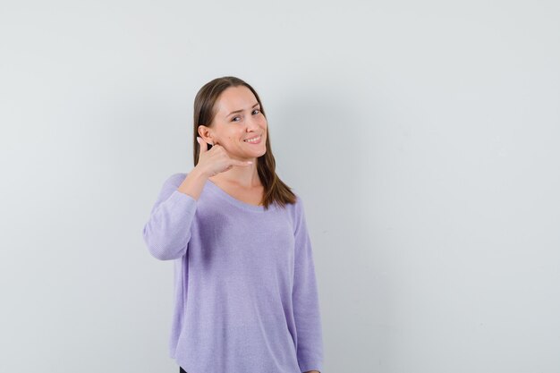 Młoda kobieta w liliowej bluzce pokazuje gest rozmowy telefonicznej i wygląda zadowolony