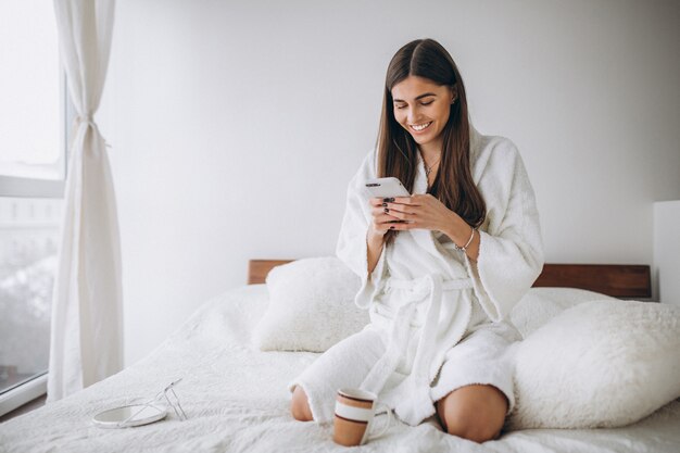 Młoda kobieta w łóżku w bathrobe z telefonem pije kawę