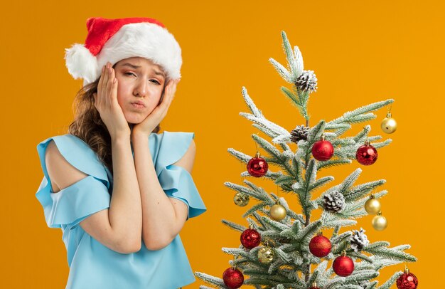 Młoda kobieta w niebieskiej bluzce i santa hat w żółtych okularach znudzona dmuchającymi policzkami, stojąca obok choinki nad pomarańczową ścianą