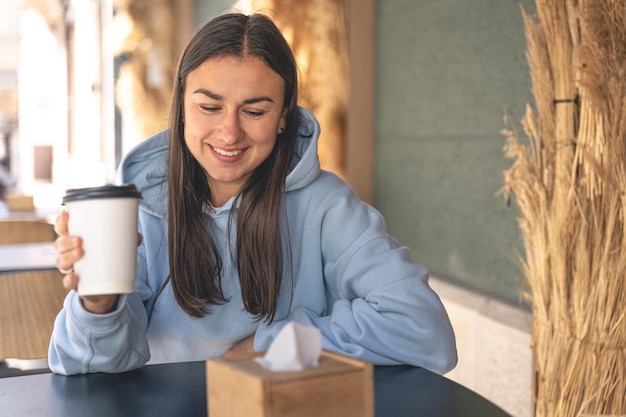 Młoda kobieta w niebieskiej bluzie z kapturem pije kawę rano w kawiarni