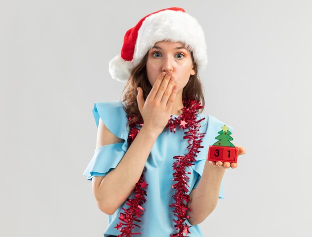Młoda kobieta w niebieskim topie i czapce mikołaja ze świecidełkiem na szyi trzymająca kostki z zabawkami z szczęśliwą datą ney roku wyglądająca na zszokowaną zakrywającą usta ręką