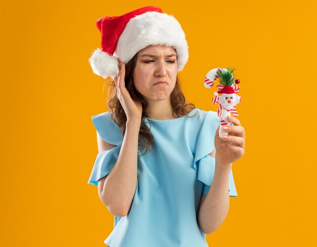 Młoda kobieta w niebieskim topie i santa hat trzyma świąteczną laskę cukierków patrząc na to zdezorientowany i niezadowolony