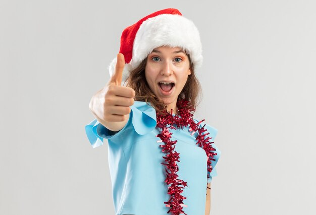 Młoda kobieta w niebieskim topie i santa hat z blichtrem na szyi wygląda na szczęśliwą i podekscytowaną pokazując kciuki do góry
