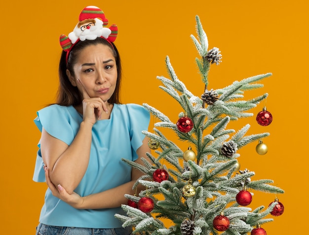 Młoda kobieta w niebieskim topie ubrana w śmieszną świąteczną obwódkę na głowie patrząc na kamerę ze sceptycznym wyrazem twarzy stojącej obok choinki na pomarańczowym tle