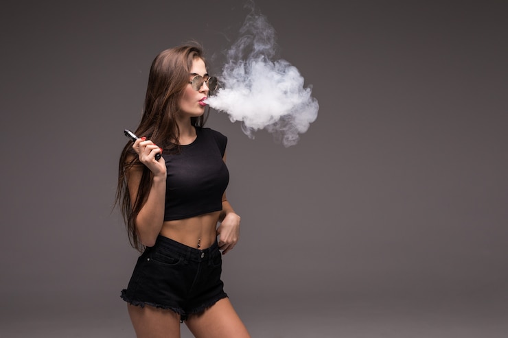 Bezpłatny plik Zdjęcie młoda kobieta w okularach przeciwsłonecznych i czarnych ubraniach vaping i dmuchanie dymem z elektronicznego papierosa na szarej ścianie