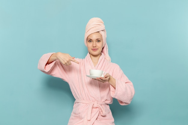 młoda kobieta w różowym szlafroku po prysznicu trzymając białą filiżankę kawy na niebieskim biurku