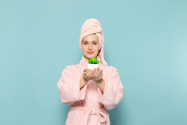 młoda kobieta w różowym szlafroku po prysznicu trzymając małą zieloną roślinę na niebiesko