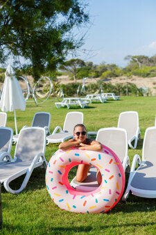 Młoda kobieta w stylowym kostiumie kąpielowym i okularach przeciwsłonecznych trzyma w dłoni nadmuchiwany pierścionek z pączkami. piękna szczęśliwa dziewczyna pozuje i bawi się w słońcu. wakacje. podróż