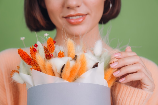 Młoda kobieta w swobodnym brzoskwiniowym swetrze na białym tle na ścianie z zielonymi oliwkami trzyma pomarańczowo-białą kompozycję kwiatów bawełny Gypsophila pszenicy i lagurusa na prezent szczęśliwy zdumiony zaskoczony