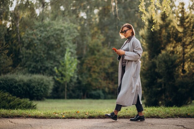 Młoda kobieta w szarym płaszczu rozmawia przez telefon w parku