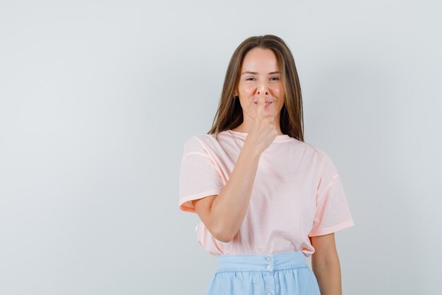 Młoda kobieta w t-shirt, spódnica dotykając nosa palcem i patrząc wesoło, widok z przodu.