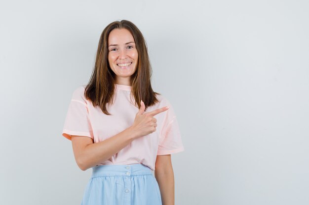 Młoda kobieta w t-shirt, spódnica, wskazując na bok i patrząc pozytywnie, widok z przodu.