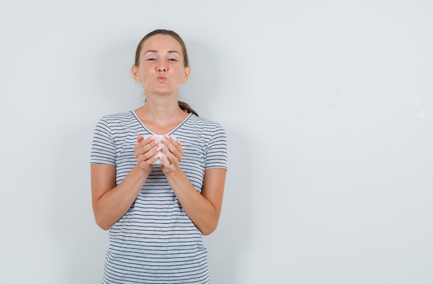 Młoda Kobieta W T-shirt, Trzymając Filiżankę Herbaty Z Zaokrąglonymi Ustami, Widok Z Przodu.