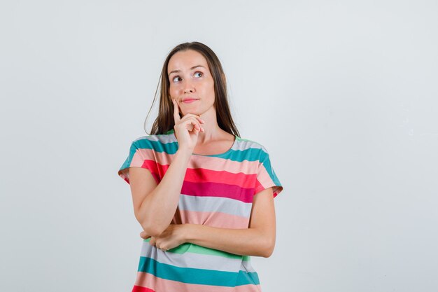 Młoda kobieta w t-shirt, trzymając palec na policzku i patrząc zamyślony, widok z przodu.