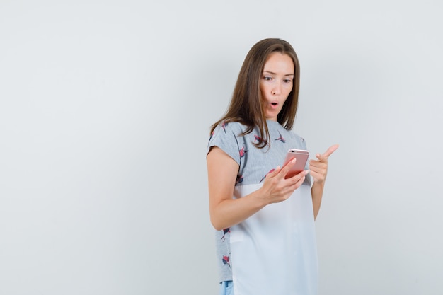 Młoda Kobieta W T-shirt, Wskazując Na Telefon Komórkowy I Patrząc Zdumiony, Przedni Widok.