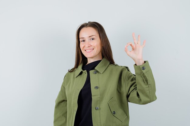 Młoda kobieta w zielonej kurtce pokazując ok gest i patrząc optymistycznie, widok z przodu.