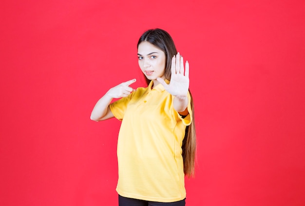 Młoda kobieta w żółtej koszuli stoi na czerwonej ścianie i coś zatrzymuje