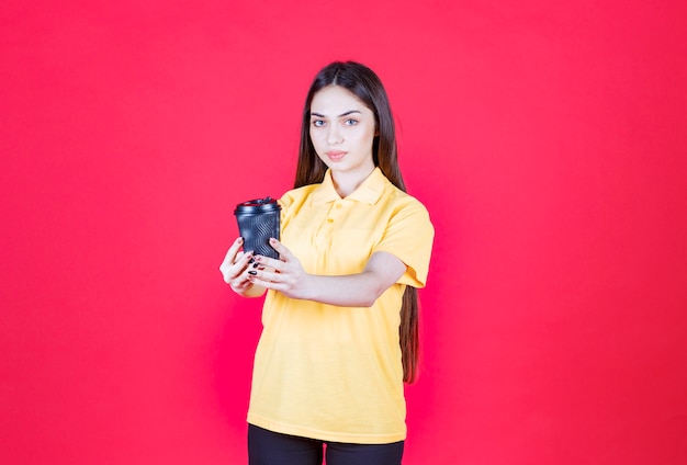Młoda kobieta w żółtej koszuli trzyma czarną jednorazową filiżankę kawy i zaprasza swojego partnera do dzielenia się