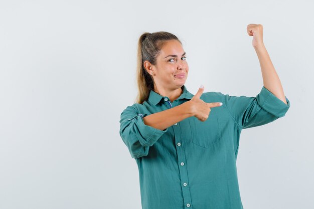 Bezpłatne zdjęcie młoda kobieta wskazując mięśnie palcem wskazującym w zielonej bluzce i wygląda atrakcyjnie