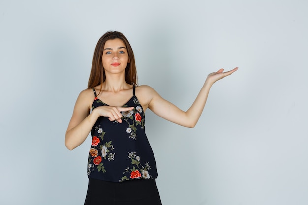 Młoda kobieta, wskazując na bok, rozkładając dłoń w kwiatowy top i wyglądająca atrakcyjnie