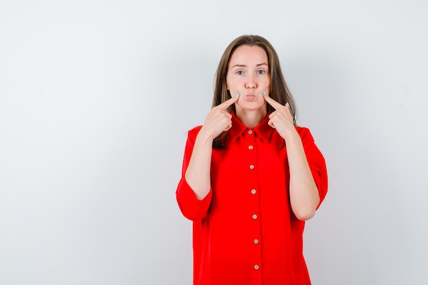 Młoda kobieta, wskazując na swoje policzki, wydębione usta w czerwonej bluzce i milcząca. przedni widok.