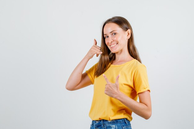 Młoda kobieta, wskazując na telefon gest w t-shirt, spodenki i patrząc zadowolony. przedni widok.