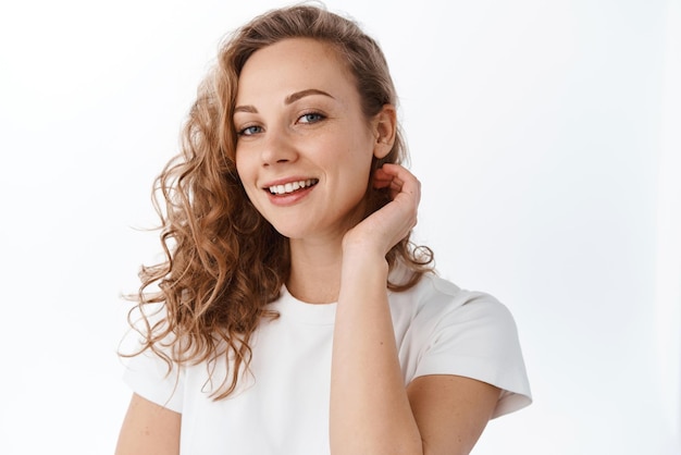 Młoda kobieta z kręconymi blond włosami założonymi za ucho i uśmiechniętymi szczęśliwie przed kamerą wygląda naturalnie i zrelaksowana, stojąc na białym tle