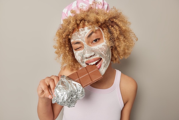 Bezpłatne zdjęcie młoda kobieta z kręconymi włosami, która gryzie słodką czekoladę mruga okiem, stosuje maskę kosmetyczną do leczenia skóry nosi kapelusz kąpielowy i dorywczą koszulkę na białym tle nad szarym tłem binge eating concept