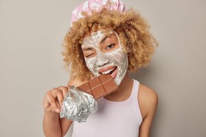 Młoda kobieta z kręconymi włosami, która gryzie słodką czekoladę mruga okiem, stosuje maskę kosmetyczną do leczenia skóry nosi kapelusz kąpielowy i dorywczą koszulkę na białym tle nad szarym tłem binge eating concept