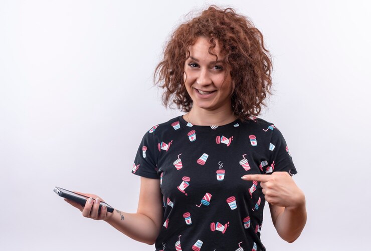 Bezpłatny plik Zdjęcie młoda kobieta z krótkie kręcone włosy trzymając smartfon wskazując palcem na to uśmiechnięty wesoło stojąc na białej ścianie