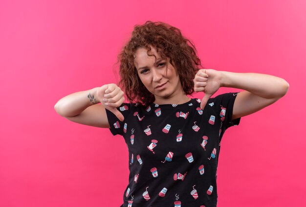 Młoda kobieta z krótkimi kręconymi włosami, niezadowolona, pokazująca kciuki w dół obiema rękami stojącymi na różowej ścianie