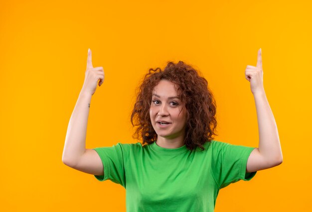 Młoda kobieta z krótkimi kręconymi włosami w zielonej koszulce patrząc uśmiechnięty, wskazując palcami, mając świetny pomysł na stojąco