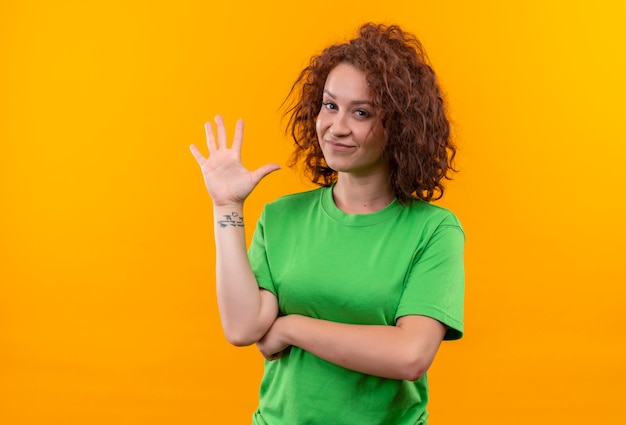 Młoda kobieta z krótkimi kręconymi włosami w zielonej koszulce uśmiechnięty macha ręką stojącą na pomarańczowej ścianie