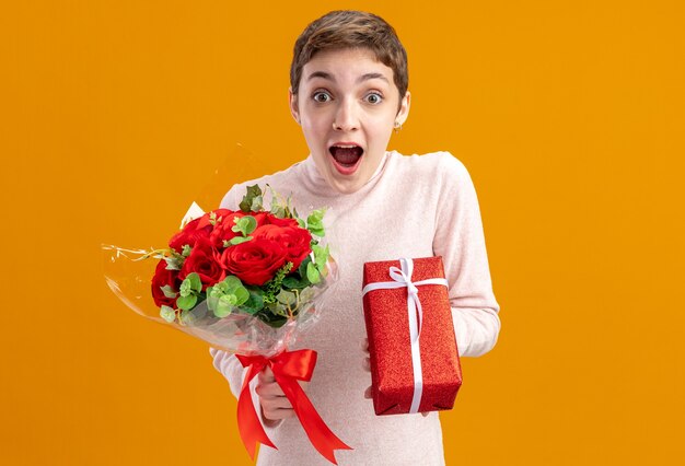 młoda kobieta z krótkimi włosami trzymająca bukiet czerwonych róż i prezent patrząc w kamerę zdumiona i zdziwiona koncepcja walentynek stojąca nad pomarańczową ścianą