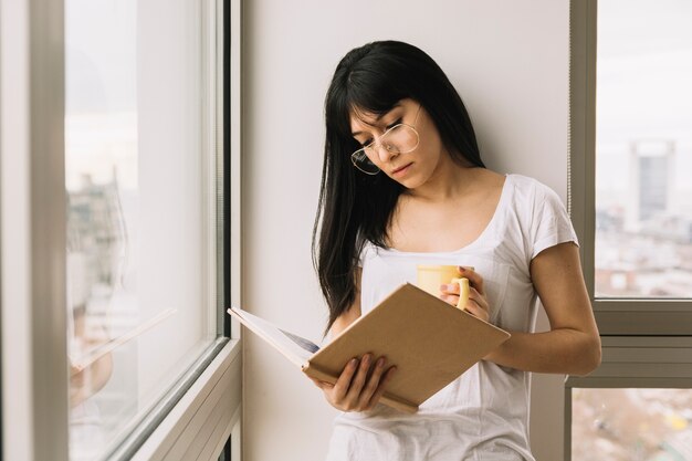 Młoda kobieta z napojem czyta blisko okno