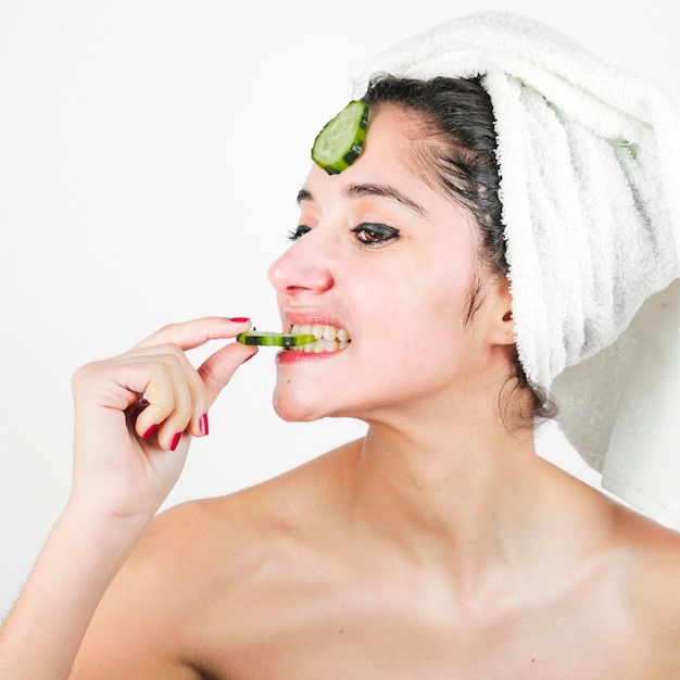 Młoda kobieta z ręcznikiem owiniętym wokół głowy jedzenia plasterka ogórka