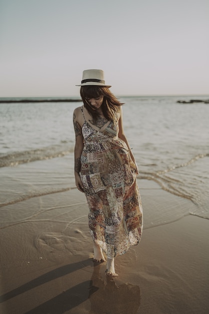 Bezpłatne zdjęcie młoda kobieta z tatuażami ubrana w sukienkę i słomkowy kapelusz na zamazanym oceanie
