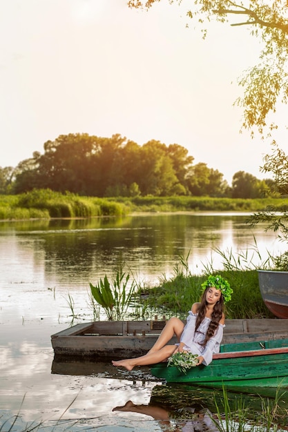 Młoda kobieta z wieńcem kwiatów na głowie, relaks na łodzi na rzece o zachodzie słońca. Piękne ciało i twarz. Fotografia artystyczna fantasy. Koncepcja kobiecego piękna, odpoczynek na wsi