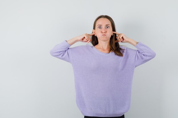Młoda kobieta zatykająca uszy palcami w liliowej bluzce i wyglądająca na znudzoną. przedni widok.
