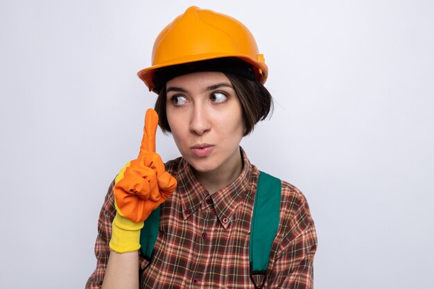 Młoda konstruktorka w mundurze budowlanym i kasku ochronnym w gumowych rękawiczkach, patrząc na bok z poważną twarzą pokazującą palec wskazujący stojący nad białą ścianą