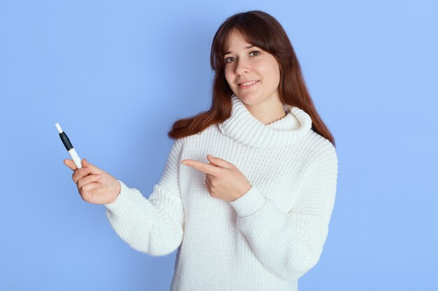 Młoda ładna ciemnowłosa kobieta wskazała na swojego elektronicznego papierosa vape z palcem wskazującym na niebiesko, uśmiechnięta urocza dziewczyna z e ciggy, ubiera się niedbale.