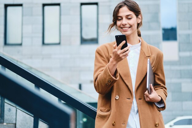 Młoda ładna dorywcza bizneswoman w płaszczu z laptopem radośnie korzystająca z telefonu komórkowego na zewnątrz