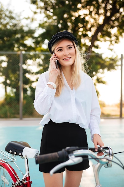 Młoda ładna Dziewczyna Z Blond Włosami W Koszuli I Szortach Stoi Z Czerwonym Rowerem Na Boisku Do Koszykówki W Parku I Rozmawia Przez Telefon Portret Uśmiechnięta Dama W Czarnej Czapce Z Daszkiem, Patrząc Na Bok