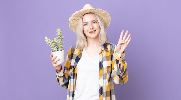 Młoda ładna kobieta albinos uśmiechnięta i wyglądająca przyjaźnie, pokazująca cyfrę cztery i trzymająca kaktusa z rośliną doniczkową